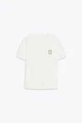 Camiseta blanca de algodón, de corte recto, cuello redondo y manga corta. Presenta un broche metálico dorado en el frente.