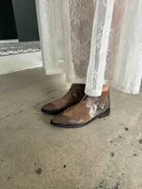 Bota corta de cuero color marrón, con taco cuadrado y punta fina. Presenta una correa con tachas pequeñas y una argolla metálica decorativa alrededor del tobillo. Cuenta con cierre lateral.