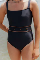 Traje de baño entero negro de escote cuadrado y tirantes anchos. Presenta un detalle de fajín ancho en la cintura con ribete marrón y botones decorativos marrones.