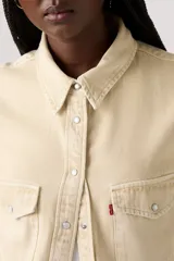 Camisa de manga larga color beige claro, estilo western, con cuello y botones a presión frontales. Presenta dos bolsillos de parche con solapa en el pecho y un pequeño tag rojo en el bolsillo derecho.