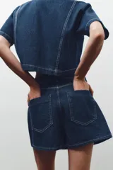 Falda pantalón corta de tiro alto confeccionada en denim azul oscuro. Presenta bolsillos delanteros y traseros de parche. El cierre es cruzado con cremallera oculta y botón metálico.