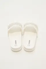 Sandalias tipo slider blancas, con tira ancha decorada con trenzado de hilo beige y plateado, tachas piramidales plateadas y flecos blancos en los bordes.