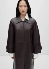 Abrigo largo oversize de cuero ovino color marrón oscuro, con cuello camisero, cierre frontal oculto con cremallera, mangas largas con puños abotonados, forro interior y dos bolsillos laterales.