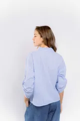 Camisa celeste a rayas verticales blancas, de corte holgado y mangas 3/4 con trabilla. Presenta cuello clásico y cierre frontal con botones.