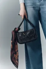 Cartera bandolera de cuero vacuno color negro, con correa trenzada y solapa con diseño calado y tachas. Incluye pañuelo estampado atado al asa.