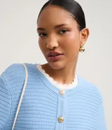 Cardigan de punto blanco, con cuello redondo, manga larga y cierre frontal de botones dorados. Posee dos bolsillos cuadrados en la parte delantera inferior.