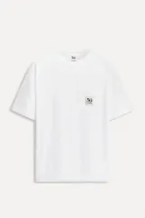 Camiseta blanca de algodón de manga corta y cuello redondo. Tiene un bolsillo de parche en el pecho con un logo bordado conmemorativo del 50 aniversario.