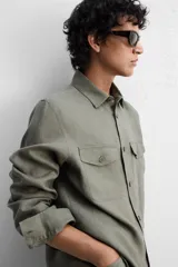 Sobrecamisa verde militar de lino, corte relaxed fit, con cuello solapa, manga larga con puños abotonados, bolsillos de parche con solapa en el pecho, aberturas laterales en el bajo y cierre frontal abotonado.