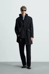 Parka negra de tejido técnico ligero, corte regular, cuello alto con capucha ajustable, manga larga con puños ajustables, cintura ajustable con cordón interno, bolsillos de solapa en la cadera, abertura en la espalda y cierre frontal con cremallera oculta y botones a presión.