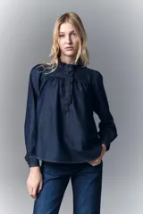 Camisa de denim azul oscuro con cuello mao, corte recto, mangas largas con puños abotonados, cierre frontal con botones a presión y detalle de volados en el cuello y la pechera.