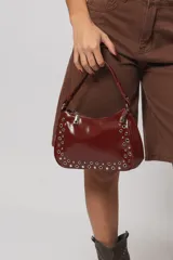 Cartera de mano color bordó charolada con correa corta y detalles de ojalillos metálicos plateados en los bordes.