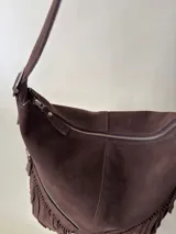 Bolso tipo hobo color tostado, hecho de cuero con flecos largos en el frente y dos bolsillos con cierre. Tiene una manija regulable con hebillas metálicas.