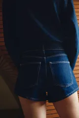 Short de jean azul oscuro, tiro alto, con cierre y doble botón metálico dorado. Presenta corte holgado y costuras visibles en hilo contrastante claro.
