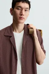 Camisa de manga corta con cuello bowling y calce relajado. Presenta un tejido con textura estructurada y efecto lavado, con cierre frontal de botones al tono.