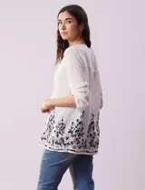 Blusa blanca de gasa bambula con cuello a la base, escote en V y pliegues. Tiene apertura central con botones, mangas ¾ con terminación fruncida y ruedo con detalles bordados en negro.