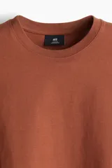 Remera marrón de algodón pesado, corte holgado, cuello redondo ribeteado, hombros caídos y bajo recto.