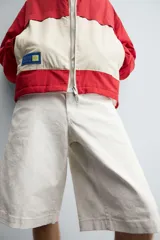 Bermuda de denim color blanco roto con efecto encerado, corte amplio (baggy fit) y cinco bolsillos. Presenta cierre frontal con cremallera y botón.