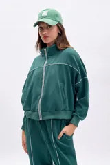 Campera deportiva de tejido de punto color verde oscuro, con cierre frontal plateado y cuello alto. Presenta vivos blancos que delinean el canesú y los puños y cintura son de elástico.