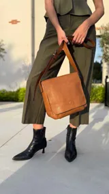 Bandolera de hombro color gris claro, con solapa frontal y correa ajustable. Presenta un diseño simple y práctico, ideal para uso diario.