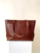 Cartera tipo tote de cuero graneado color negro, con textura uniforme y suave al tacto. Tiene manijas de 55 cm, bolsillo frontal con broche imantado, bolsillo trasero con cierre metálico, cierre principal metálico, forro interior y tres bolsillos internos (uno con cierre, uno plaqué y uno tipo sobre con solapa y botón).