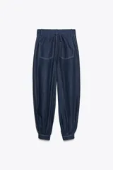 Pantalón bombacho de denim azul oscuro, tiro medio, con trabillas en la cintura, bolsillos delanteros y traseros, pespuntes marcados a contraste, bajo abullonado con puño y cierre frontal con cremallera, botón y ganchos metálicos.
