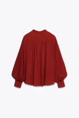 Blusa de cuello subido color ladrillo, confeccionada en hilatura de viscosa 100%. Manga larga con pliegues y abullonada. Detalle aplicación de lazo en cuello ajustable con lazada. Cierre frontal con botones.