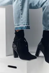Botas de caña media de gamuza negra, con taco cuadrado y punta fina. Presenta tiras terminadas con borlas que se cruzan y atan alrededor de la caña.