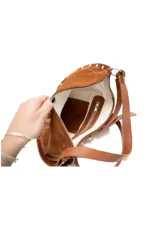 Cartera de gamuza color marrón, de tamaño mediano y forma curva. Posee una correa ajustable y detalles de pequeñas tachuelas metálicas en el borde inferior.