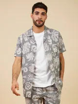 Camisa manga corta gris con estampado de hojas y flores en blanco, con cuello tipo cubano y bolsillo de parche en el pecho.