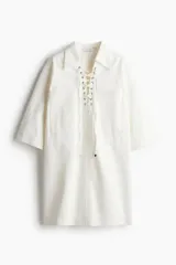 Vestido corto de denim color blanco, con cuello camisero, mangas cortas, dos bolsillos frontales y ajuste con cordones cruzados en el escote.
