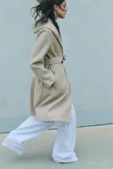 Abrigo largo color beige con capucha, cuello con solapa, manga larga, bolsillos laterales y cinturón lazada en el mismo tejido. Cierre frontal cruzado con botones.