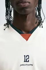 Camiseta blanca de manga larga con cuello en pico. Tiene el número 12 bordado en el pecho y dos franjas horizontales en color azul marino en cada manga.