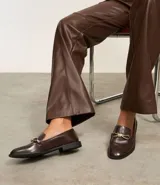 Zapato mocasín de cuero color marrón oscuro con detalle metálico dorado en la parte delantera.