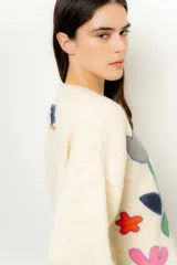 Sweater de lana color crudo con cuello redondo y mangas largas. Presenta apliques de tela cosidos con forma de flores y figuras geométricas en tonos gris, azul, verde, amarillo y rosa.