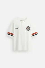 Camiseta blanca de corte holgado con cuello redondo y cierre frontal de botonadura oculta. Tiene mangas cortas con rayas horizontales en marrón, verde y azul. Presenta un parche bordado en el pecho y texto bordado en el frente.