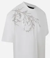 Remera de algodón color crema, corte boxy, cuello redondo y manga corta. Presenta un bordado floral en el lateral superior izquierdo.
