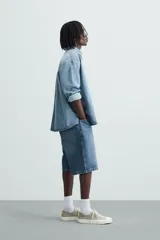 Bermuda de denim de algodón, color gris claro lavado, de corte ancho (baggy fit) y largo hasta la rodilla. Presenta cinco bolsillos y cierre frontal con cremallera y botón.