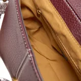 Bandolera pequeña de material sintético color bordó, con textura granulada. Presenta una solapa frontal con costuras decorativas blancas y un logo metálico con las letras "XL". Incluye una correa ancha de tela con diseño a rayas blancas y marrones, ajustable y desmontable, y herrajes metálicos dorados.