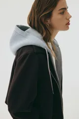 Campera negra de tejido suave efecto neopreno, con cuello y cierre frontal. Presenta un diseño de doble capa simulado con una capucha de tela gris claro que asoma por debajo del cuello de la campera.