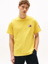 Remera amarilla de algodón con corte amplio, cuello redondo y hombros caídos. Presenta un logo pequeño de Tommy Hilfiger en el pecho, un logo en la manga y un logo grande en la espalda. Pertenece a la colección Hilfiger Sailing.
