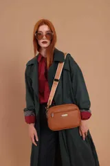 Bandolera color marrón de cuero sintético con textura granulada, tres cierres principales, cierre delantero y bolsillo trasero. Incluye correa ajustable de tela beige con franja blanca.