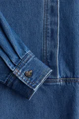 Blusa azul de denim con cuello redondo, tapeta con botones ocultos y mangas largas con puños abotonados.