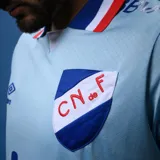 Camiseta de fútbol Umbro del Club Nacional de Football, modelo Away 3. Diseño celeste con detalles en azul y rojo en cuello y mangas. Incluye logo de Umbro, escudo del club y sponsors.