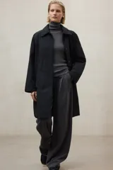 Chaqueta larga de color blanco roto, con cuello camisero y cierre frontal de broches. Presenta un corte holgado y bolsillos laterales.