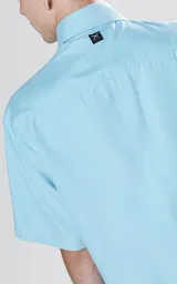 Camisa celeste de corte holgado, con cuello clásico, cierre frontal con botones, mangas cortas y cuatro bolsillos tipo cargo con solapa en el frente.