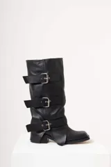 Bota de cuero negra con caña alta y tres correas con hebillas plateadas. Tiene suela de goma y taco bajo cuadrado.