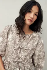 Blusa estampada color beige y blanco, con mangas abullonadas, escote redondo y lazos frontales.