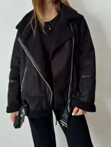 Campera negra estilo aviador, de gamuza sintética con interior de piel sintética. Tiene cuello solapa con hebillas, cierre cruzado con cremallera y cinturón con hebilla en la cintura.