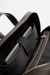 Mochila marrón de cuero sintético con textura grabada. Tiene un compartimento principal con cierre de cremallera, un bolsillo interno para dispositivos electrónicos de hasta 13 pulgadas y un bolsillo interno con cremallera. En la parte delantera tiene un bolsillo con cremallera. La parte posterior es acolchada y tiene una banda de sujeción para equipaje. Tiene un asa de mano y dos asas de hombro ajustables y acolchadas.