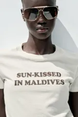 Camiseta color crema de algodón con efecto lavado, corte regular, cuello redondo, manga corta y texto marrón estampado en el frente con la frase 'Sun-Kissed in Maldives'.